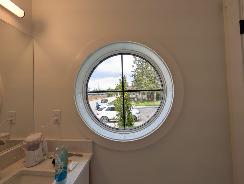 Round bathroom window with clear glass before privacy film, looking straight down onto the street and a neighbouring house in Highland Park, NW Calgary