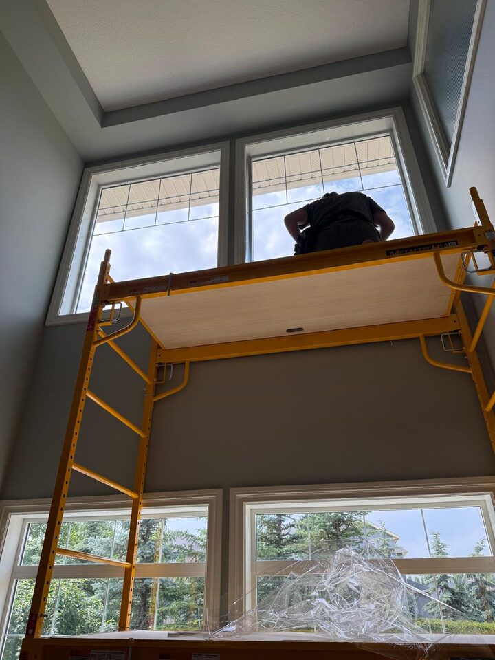 Installer on interior scaffolding applying solar window film to tall two-storey living room windows in Silverado SW Calgary